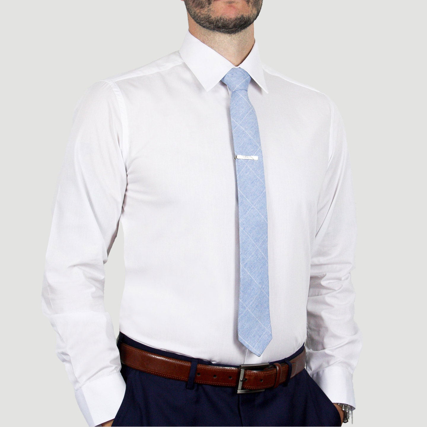 A person wearing an ARTURO Modern Fit Long Sleeve White Dress Shirt, paired with a light blue tie with a tie clip, complemented by a brown belt and dark pants made from a stretchy spandex blend, stands with hands casually tucked in their pockets.