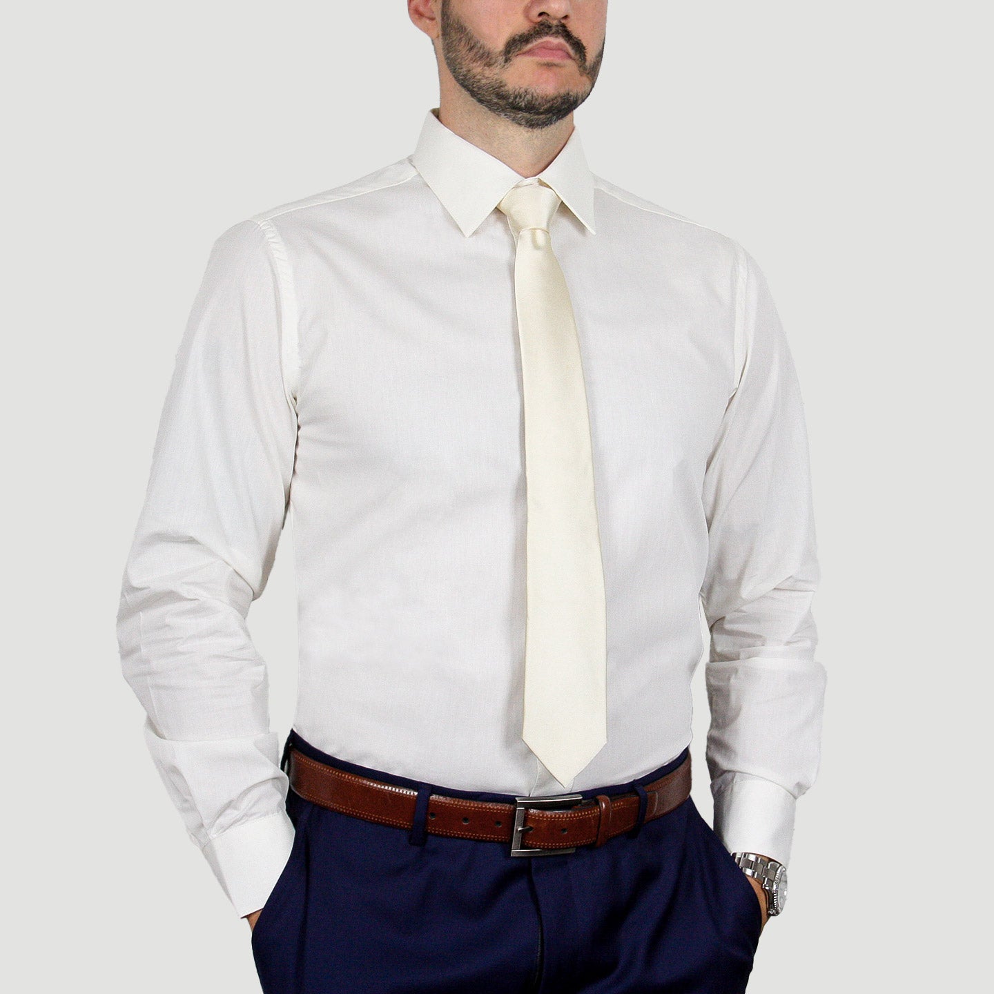 A man wearing the ARTURO Modern Fit Long Sleeve Ivory Dress Shirt, complemented by a cream tie, a brown belt, and dark polyester pants stands with hands in pockets.