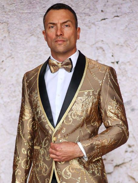 A man wearing the INSOMNIA MZS-550 CHAMPAGNE Blazer by Inserch, featuring gold embroidery and a matching bow tie, stands against a textured white background.