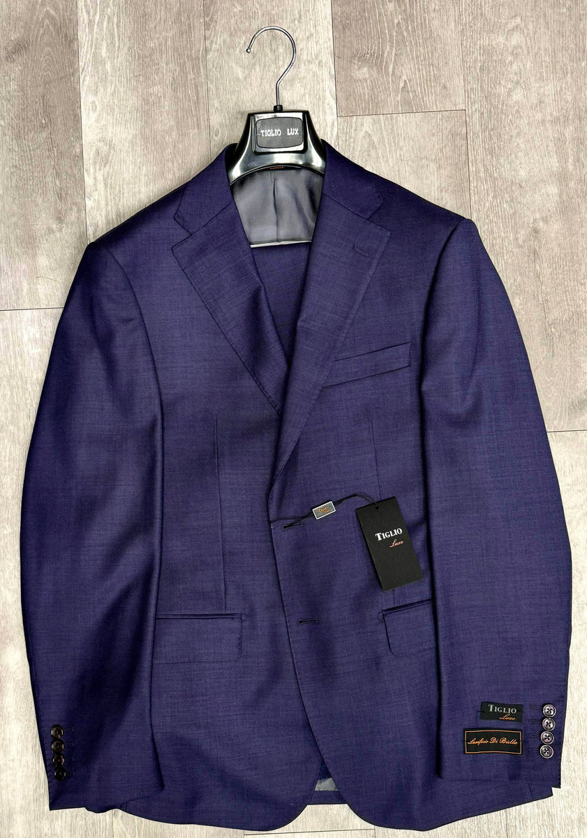 A navy blue suit jacket from the "Tiglio Luxe Dolcetto Modern Fit, Pure Wool Suit TL3124" collection, available only in sizes 38R and 42R, hangs elegantly on a hanger against a light wooden floor, showcasing the timeless sartorial traditions of the Tiglio Luxe brand.