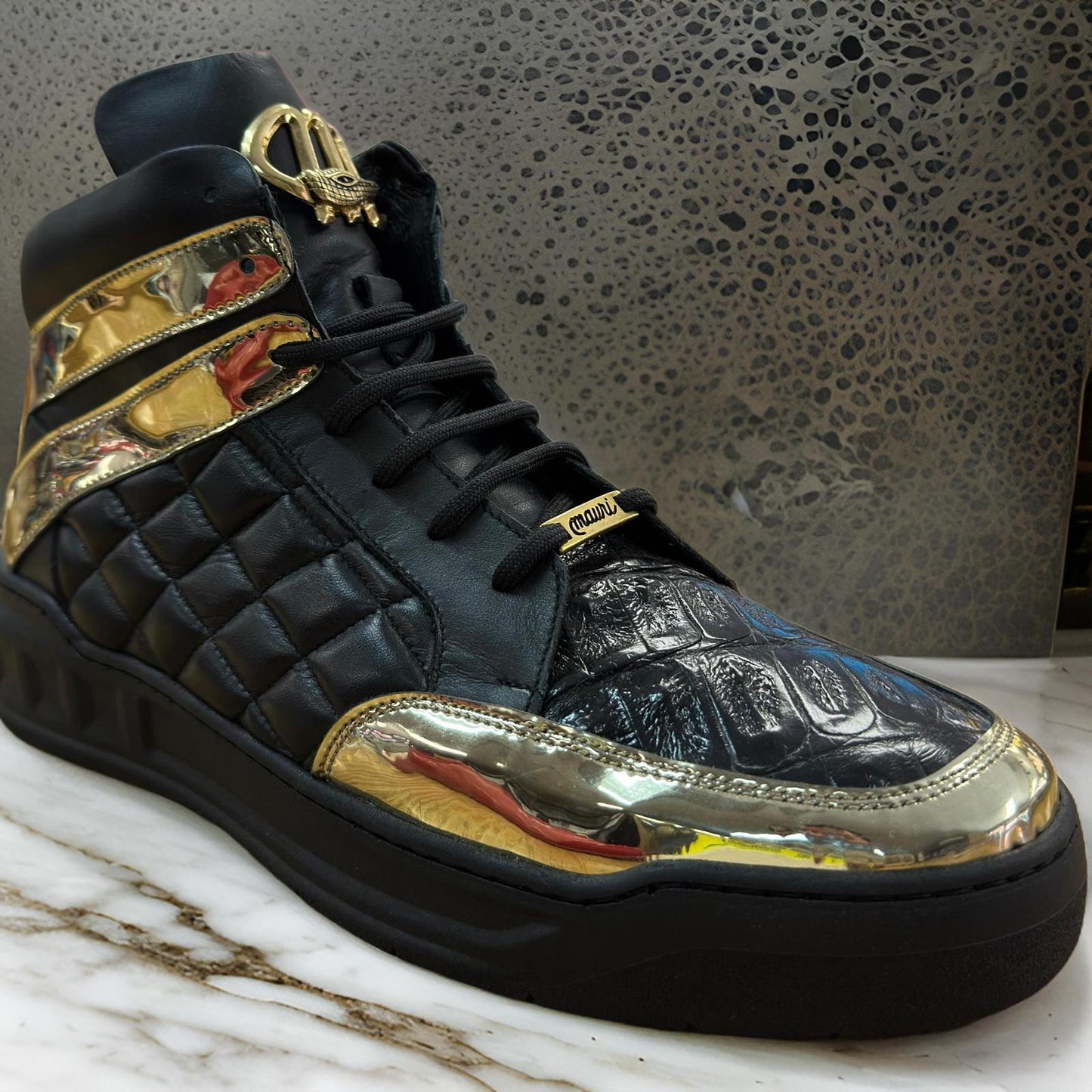 The custom-designed Mauri 8499 black and gold high-top boot, featuring a quilted texture and metallic detailing, gracefully sits on a marble surface.