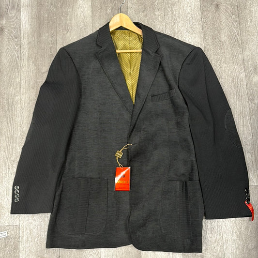 The Inserch Blazer BL506-01 in black, featuring contrasting textures and patch pockets for a refined appearance, drapes gracefully on a wooden hanger above the luxurious wooden floor. The sleeve is adorned with a bold red tag, enhancing its sophisticated appeal.