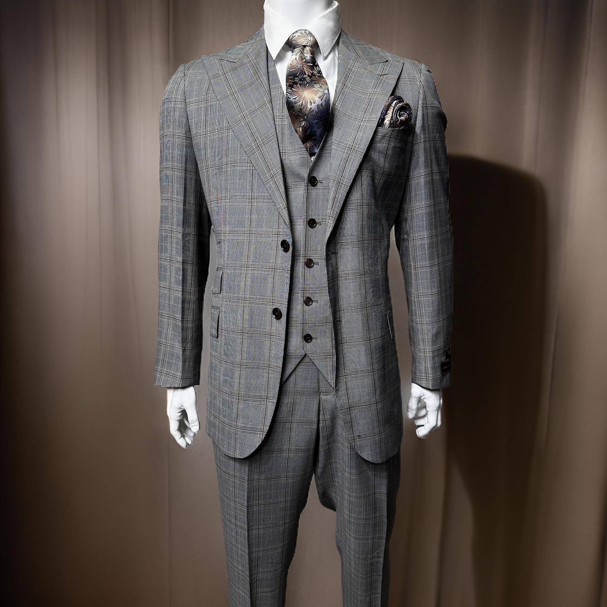 Displayed on a mannequin against a plain background is the Tiglio Luxe Prosecco Suit/Vest in grey/brown plaid from Unique Design Menswear. This exquisite three-piece suit, expertly crafted from Italian fabrics, features a dark patterned tie and pocket square, exemplifying the elegance of fine men's clothing.