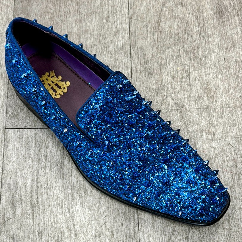 The Exclusive Formal Dress Shoe Royal Blue 6788 from UNIQUE DESIGN MENSWEAR, featuring pointed studs on the top and crafted from genuine leather with a glittery blue finish, elegantly rests on a wood-like surface.