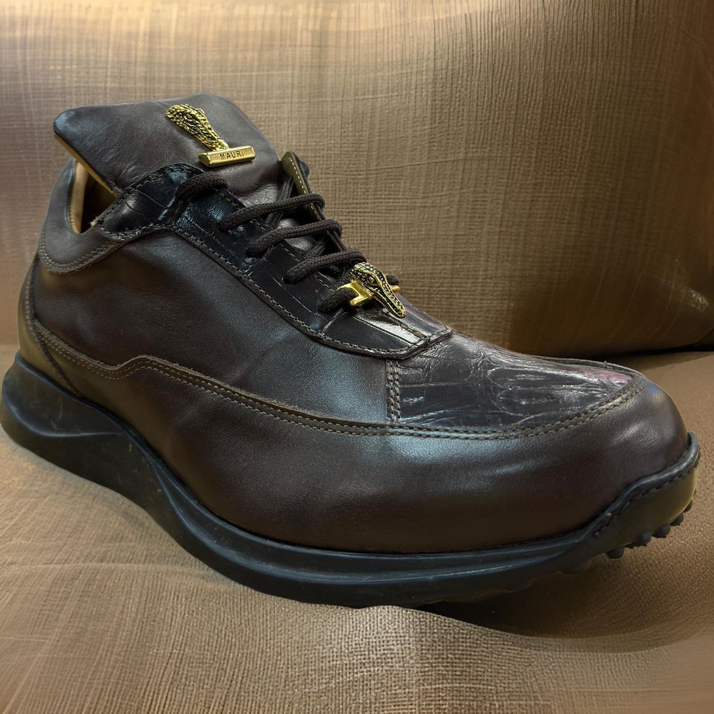 A pair of Mauri 8900 Dark Brown Shoes, featuring black laces and resting on a beige fabric background, are custom made in Italy. These sneakers showcase a textured sole and metallic accents on the tongue.