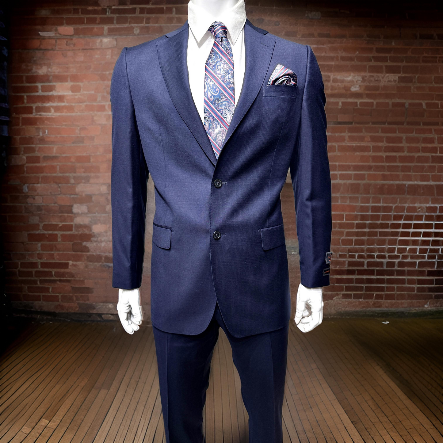 The mannequin is dressed in a Tiglio Luxe Dolcetto Modern Fit Wool Blue Tone on Tone Plaid Suit TL5237, featuring a patterned tie and pocket square, positioned against a brick wall background.