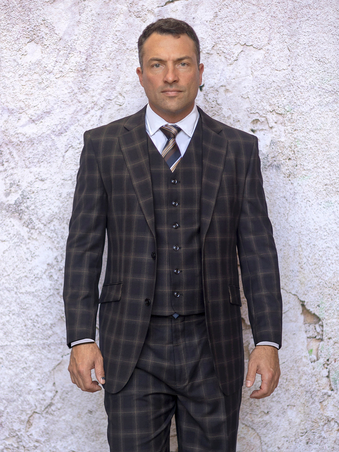 A man in an INSOMNIA ARIA 3PC Regular Fit Brown Suit from Insomnia stands against a textured wall, the pleat pants adding a touch of sophistication, with his hands relaxed by his sides as he looks at the camera.