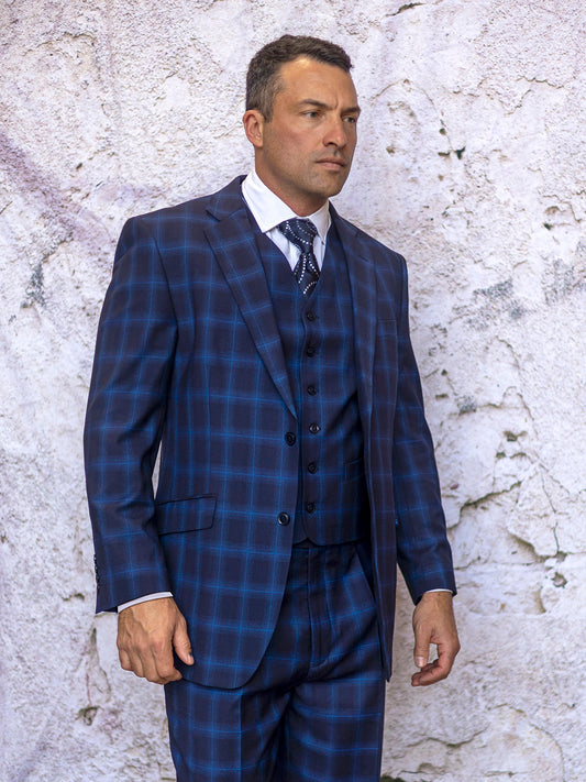 A man wearing the INSOMNIA ARIA 3PC Regular Fit Indigo Suit from Insomnia with pleated pants stands against a textured, light-colored wall.