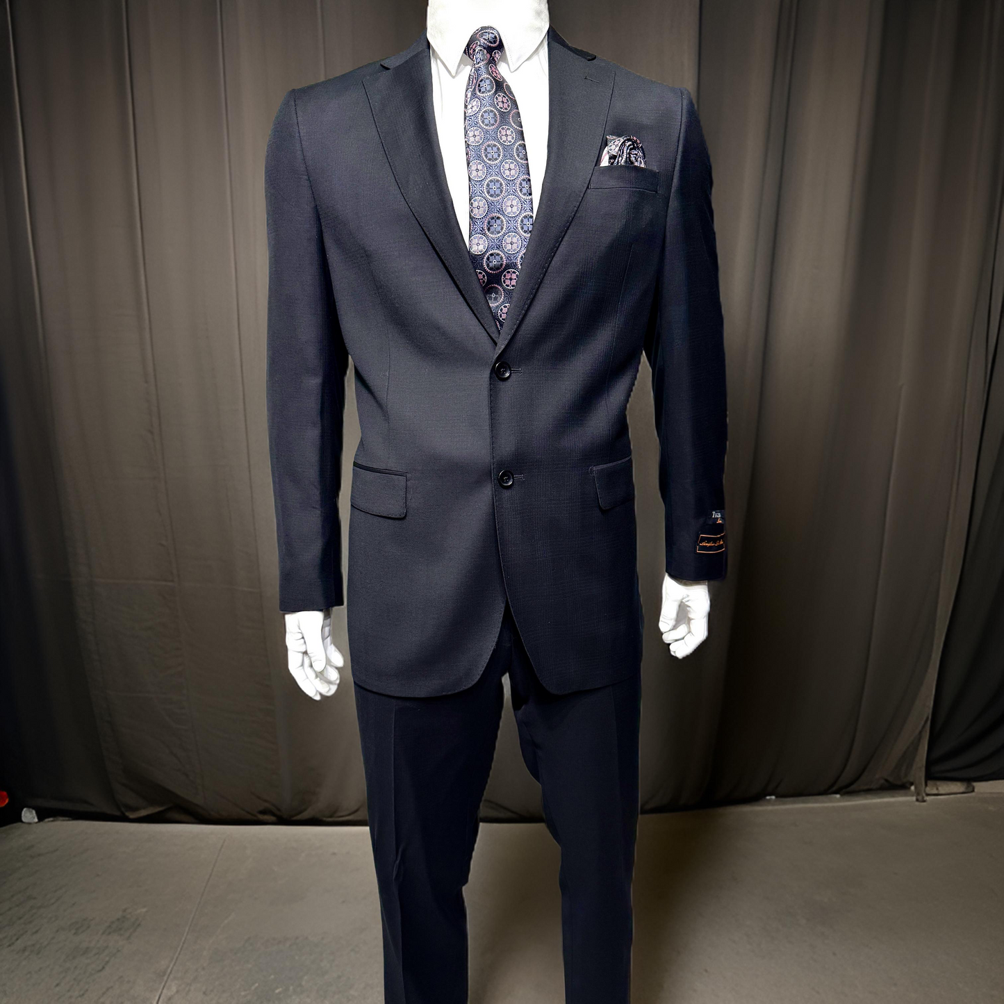 A mannequin dressed in the Tiglio Luxe Dolcetto Modern Fit Pure Wool Suit TL5236, adorned with a patterned tie and pocket square, stands elegantly against a dark curtain backdrop, highlighting the finest offerings from Tiglio Luxe in men's clothing.