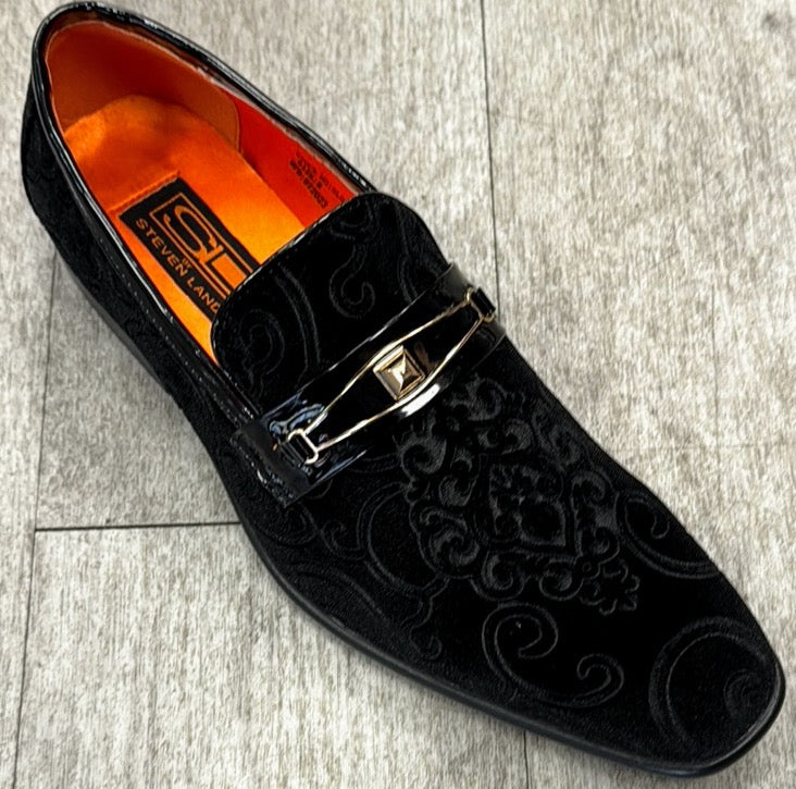 The Exclusive Formal Dress Shoe Black Paisley with Buckle SL0096 by UNIQUE DESIGN MENSWEAR is a luxurious black velvet loafer featuring embossed patterns and a gold buckle detail, elegantly displayed on a light wood floor. Expertly crafted in Italy, its interior boasts a vibrant orange lining that highlights its exquisite style and craftsmanship.