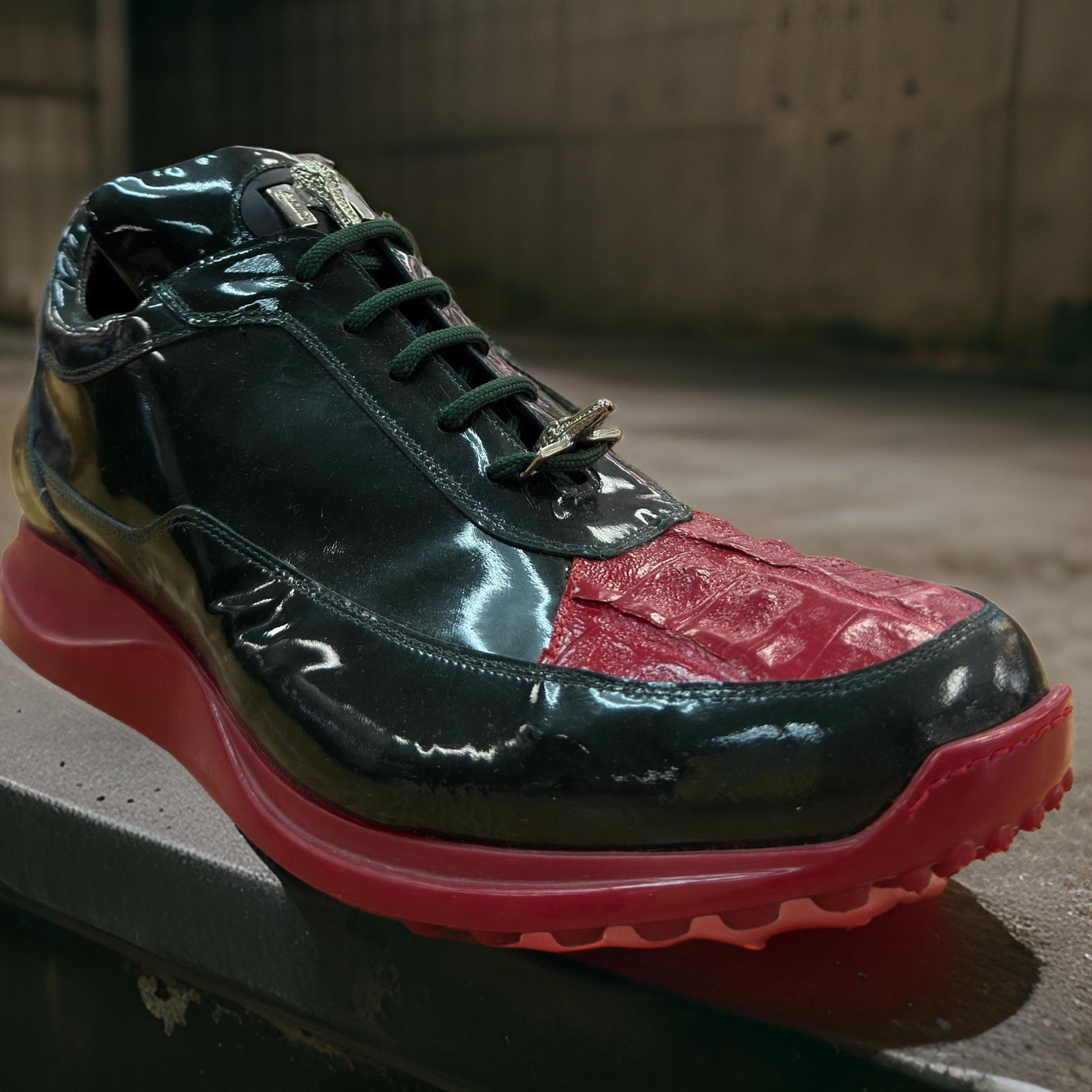 A sleek casual sneaker by Mauri, featuring glossy black and red patent leather with a metallic logo on the tongue and a red sole, rests on a concrete surface.