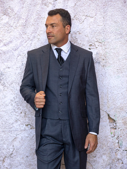 A man in an INSOMNIA CONRAD 3PC Regular Fit Charcoal Suit stands against a textured white wall, looking to the side.