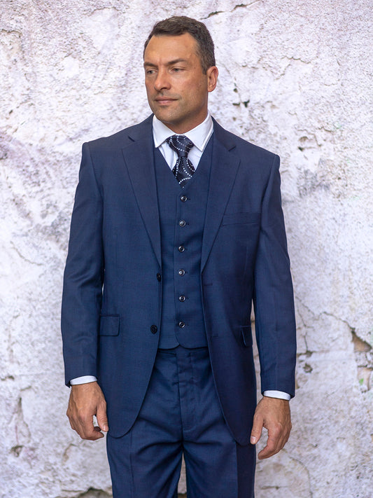 A man wearing the INSOMNIA CONRAD 3PC Regular Fit Navy Suit, complete with pleat pants and a patterned tie, stands against a textured stone wall, gazing to the side.