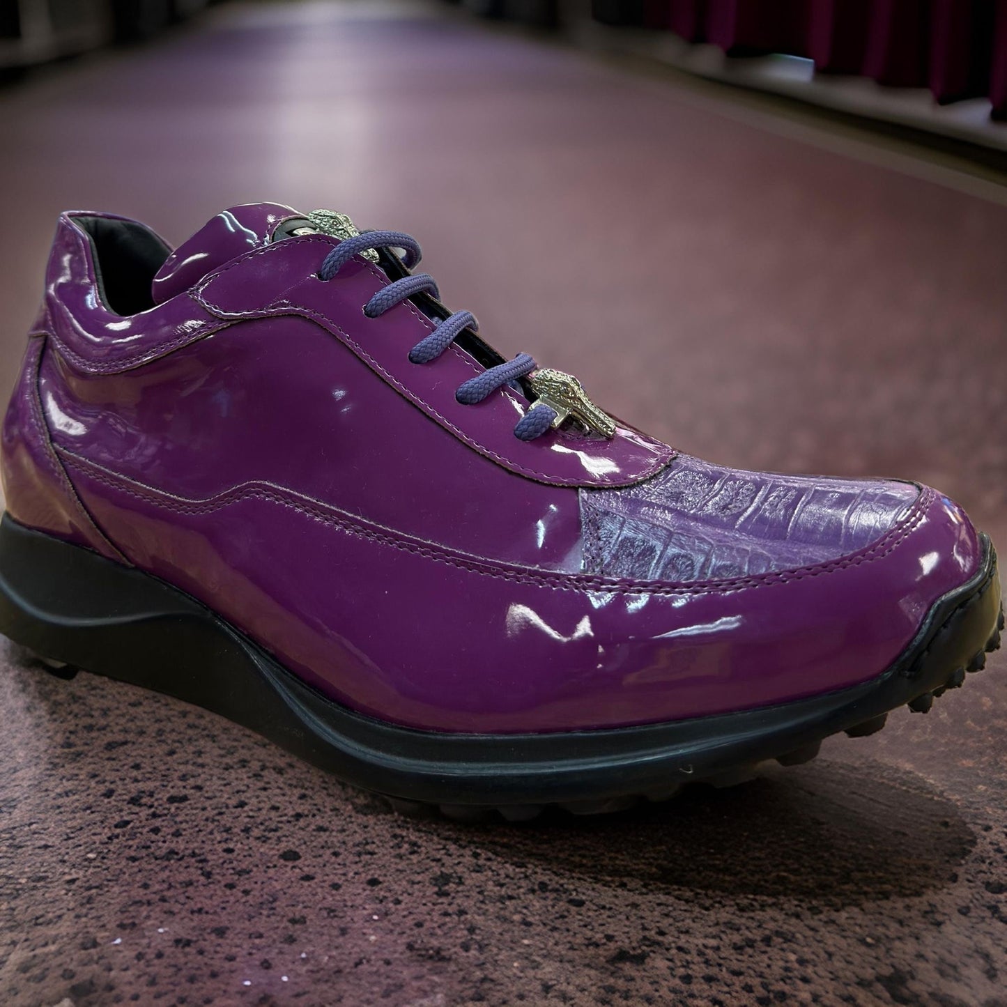 A pair of Mauri 8900 Light Purple Shoes IN STORE, featuring a glossy purple finish with black laces and a textured upper, exudes style reminiscent of the Mauri brand. These custom-made casual sneakers from Italy come with a thick black sole and are showcased on a brown speckled floor.