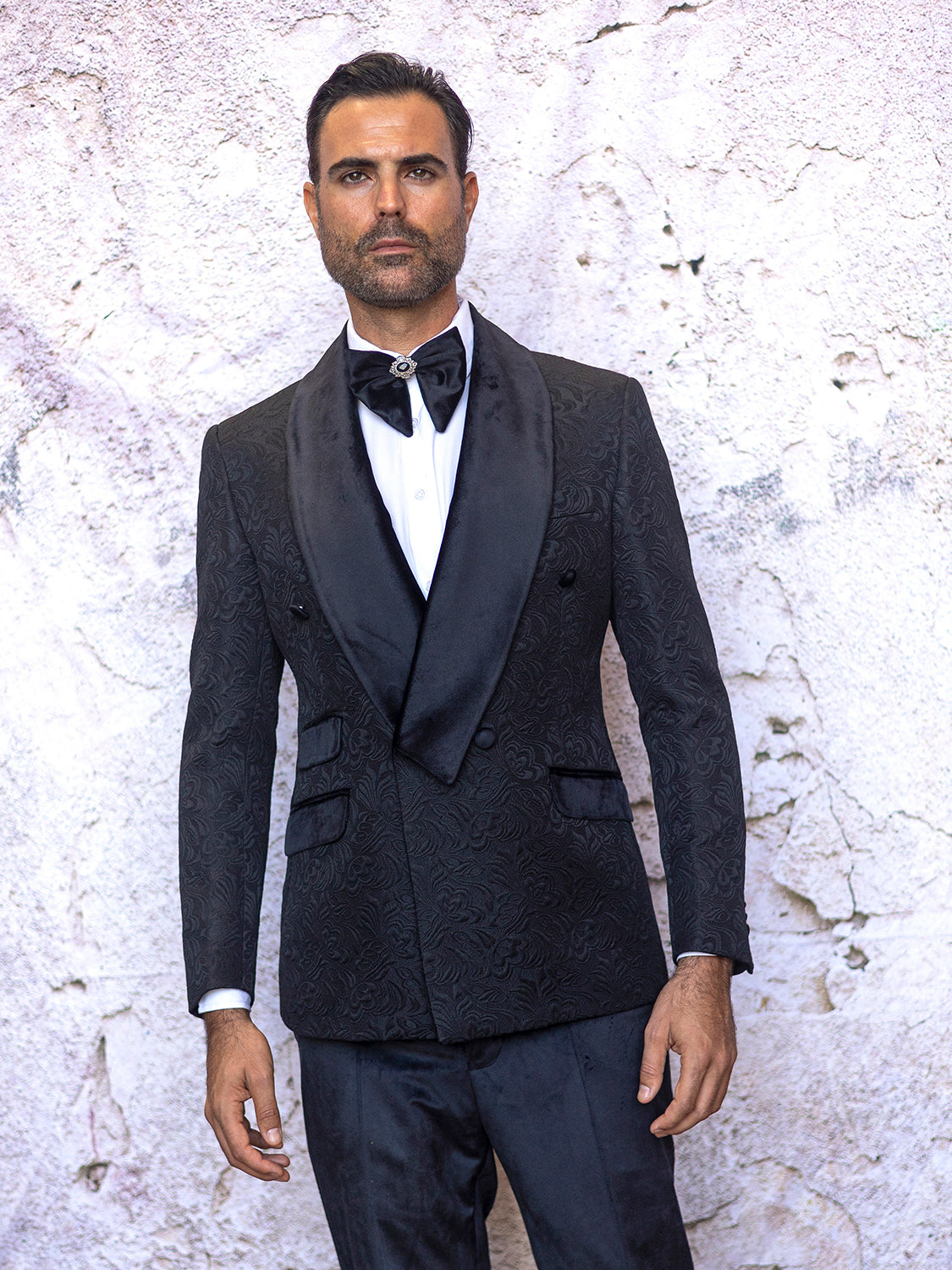 A man in an INSOMNIA DELANO 2PC Floral Pattern Slim Fit Black Suit, featuring a white shirt and velvet trim, stands against a textured cream wall, sporting a matching black bow tie.