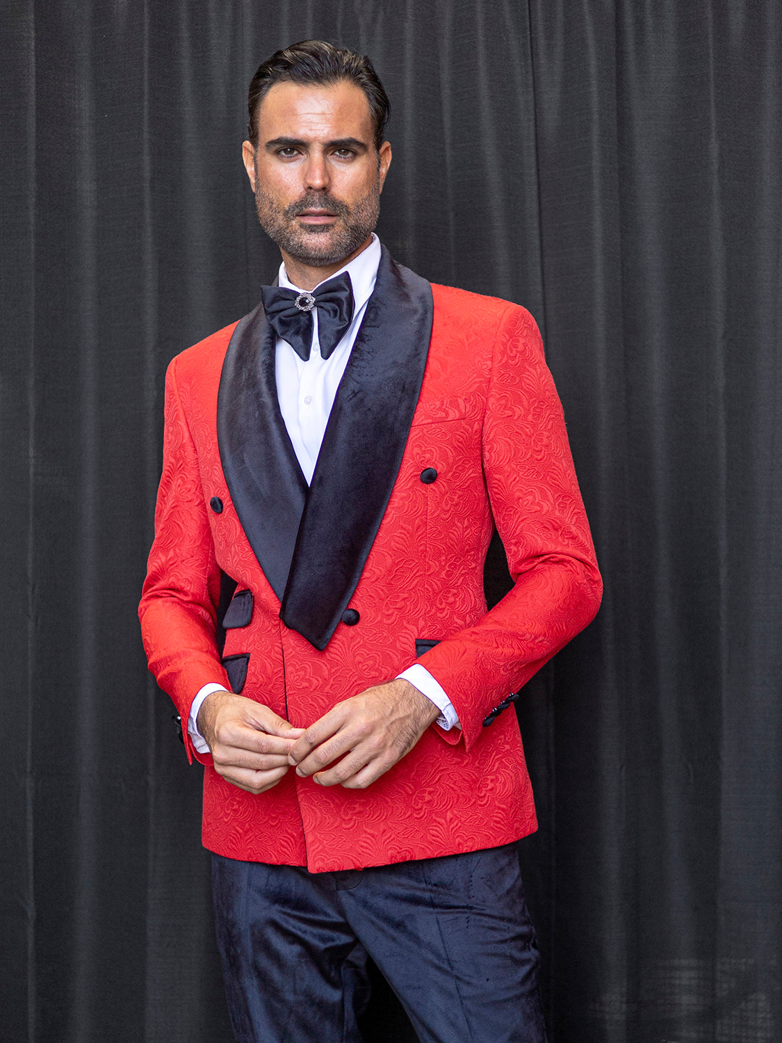 A man wearing the INSOMNIA DELANO 2PC Floral Pattern Slim Fit Red Suit by Insomnia, featuring a subtle floral print and black velvet trim, paired with a white shirt and classic black bow tie, stands in front of a dark curtain.