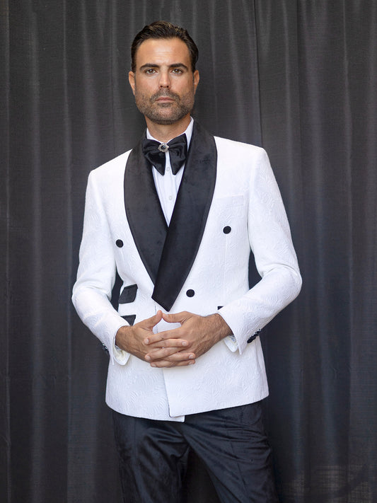 A man wearing the INSOMNIA DELANO 2PC Floral Pattern Slim Fit White Suit stands in front of a dark curtain, hands clasped.