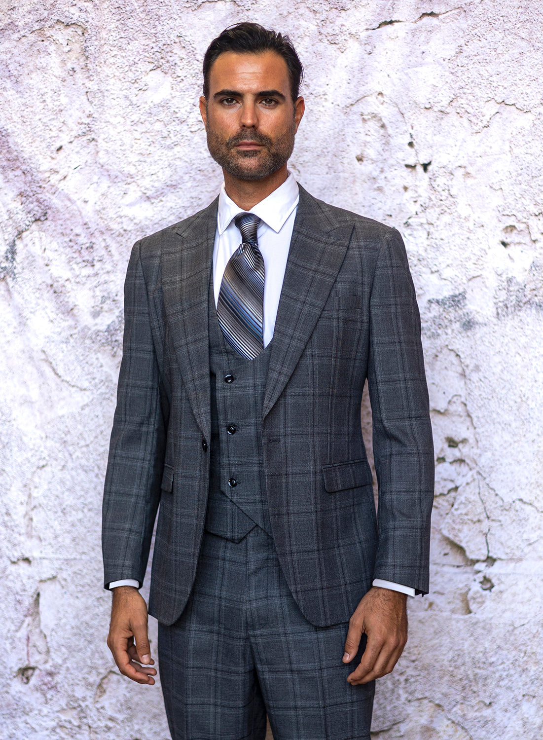 A man wearing the INSOMNIA ENCORE 3PC Tailored Fit Charcoal Suit from Insomnia stands against a textured, light-colored wall. His hair is dark, and he has a beard. He complements the suit with a striped tie and white shirt.