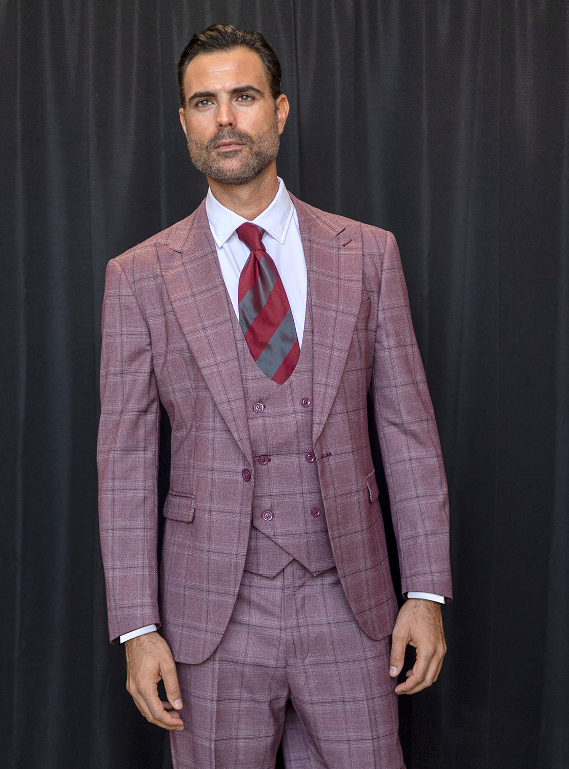 A man wearing the INSOMNIA ENCORE 3PC Tailored Fit Wine Suit from Insomnia, complemented by a red-striped tie, stands in front of a black curtain.