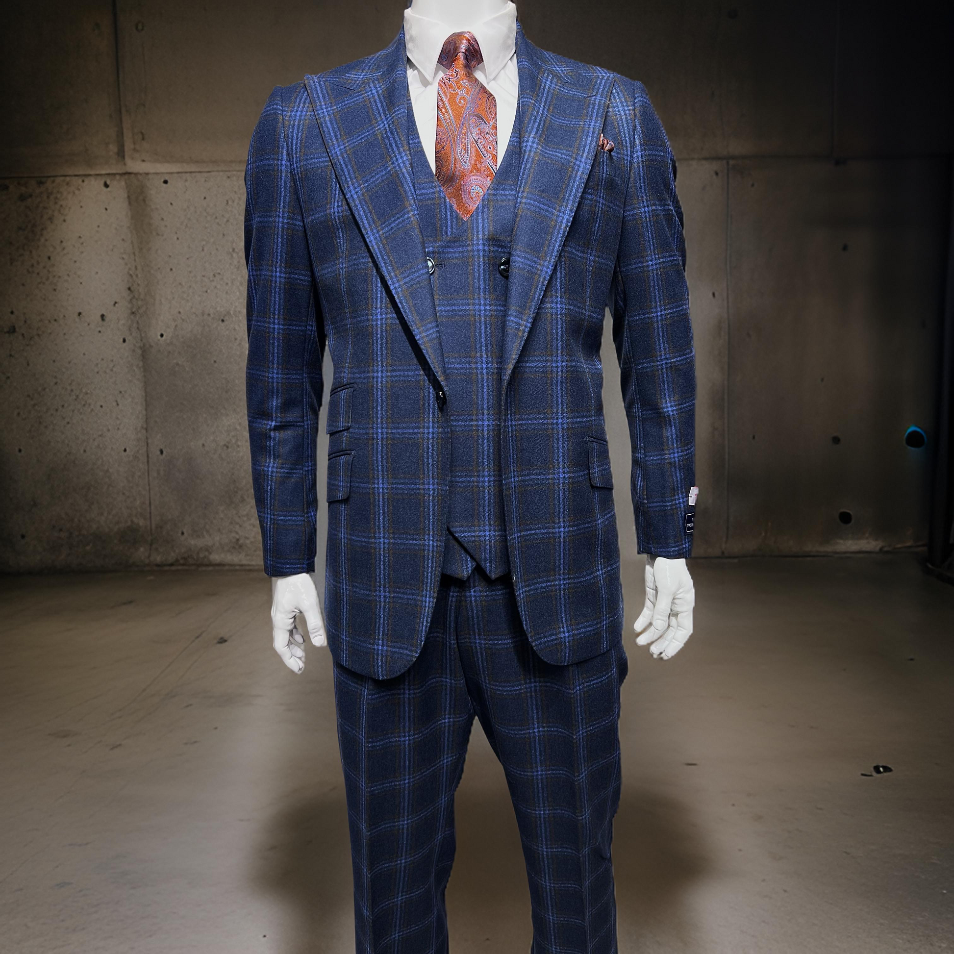 A mannequin showcases a striking Tiglio Rosso Orvietto suit in a blue and rust windowpane pattern, complemented by a colorful paisley tie against a plain, industrial backdrop. Made from 100% wool, this three-piece ensemble epitomizes elegance and quality craftsmanship.