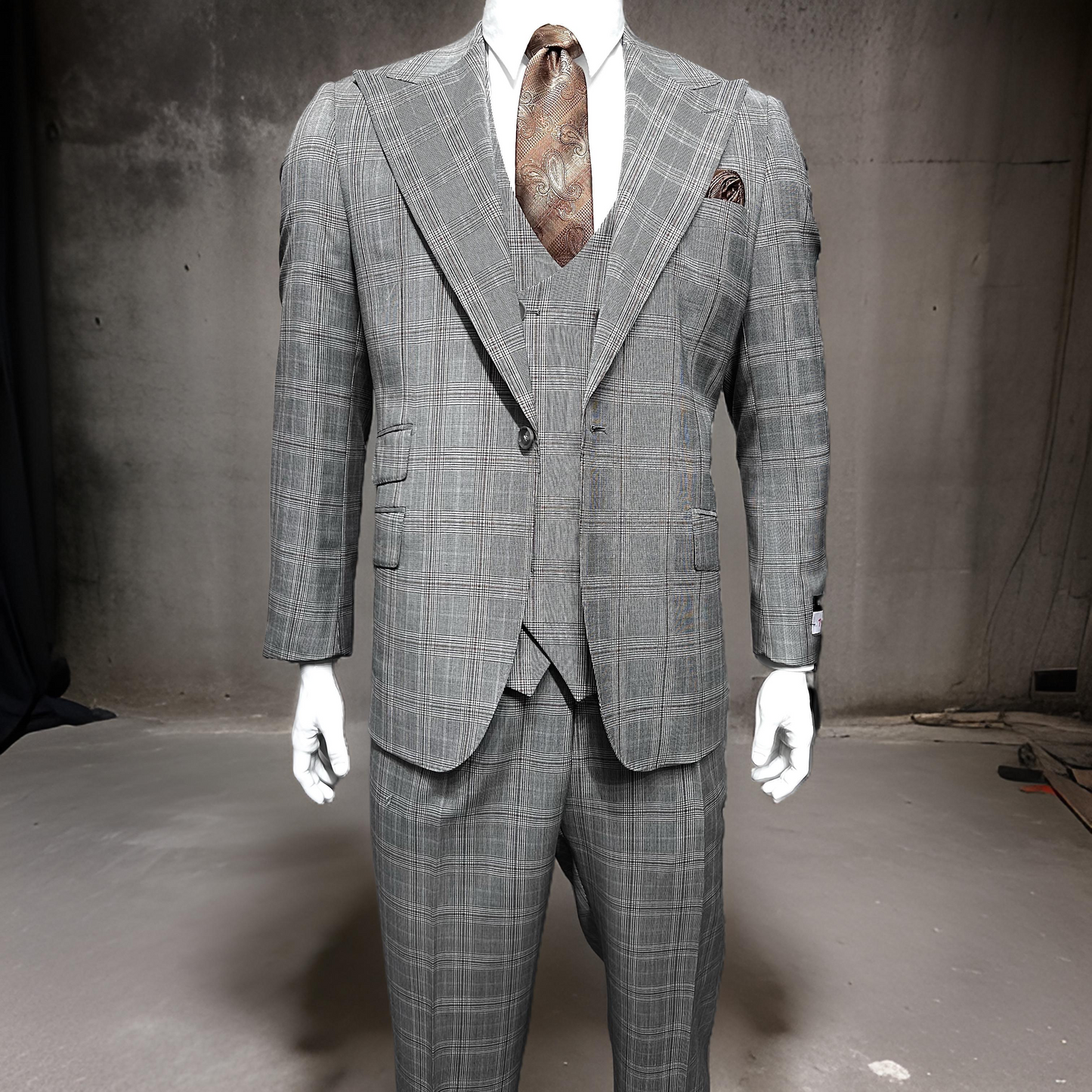 A taupe and brown plaid Tiglio Rosso Orvietto TL5203 three-piece suit, made entirely from wool, is elegantly showcased on a mannequin within a minimalist, industrial environment. This refined outfit embodies the classic elegance that is synonymous with Tiglio Rosso suits.