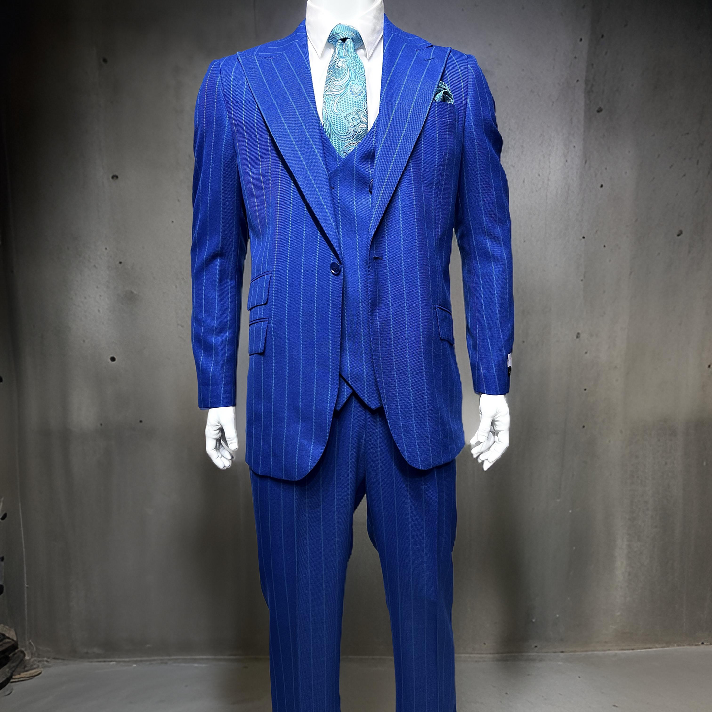 A mannequin showcases the Tiglio Rosso Orvietto Blue with Green Pinstripe TL6018 suit, crafted from 100% wool. The royal blue pinstripe three-piece ensemble includes a matching vest, crisp white shirt, and teal paisley tie, all elegantly set against a dark, plain backdrop.