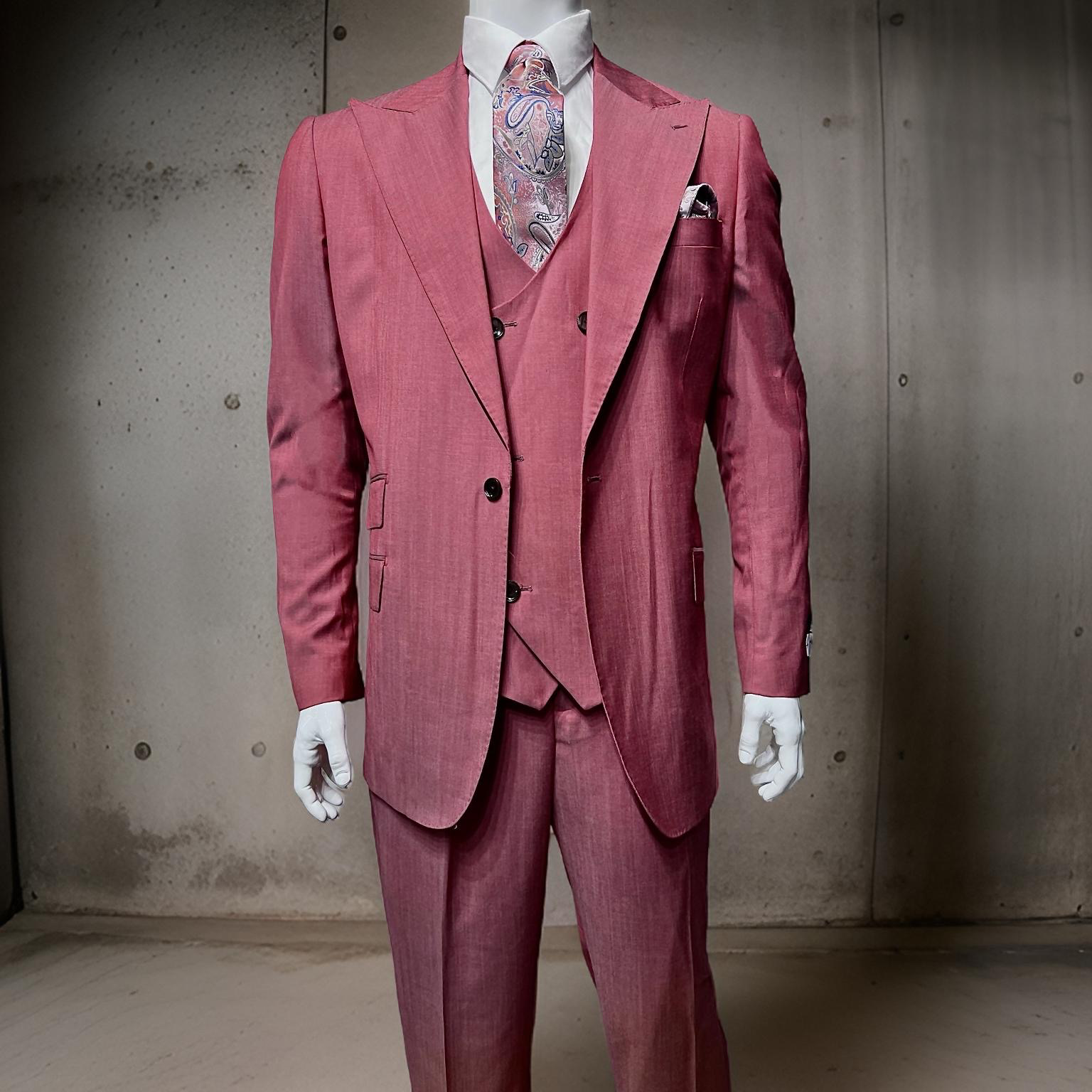 A mannequin displays the elegant Tiglio Rosso Orvietto Light Red TL4016, a vibrant three-piece pink suit crafted from 100% wool. The ensemble is accessorized with a patterned tie and pocket square, all set against a concrete wall background to highlight the sophistication of Tiglio Rosso suits.