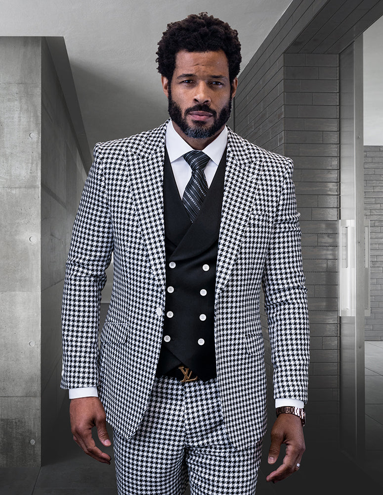 A man wearing the MASIMO-BLACK houndstooth suit by Statement Clothing, crafted from fine Italian wool and paired with a black vest and tie, stands confidently in a modern, minimalist room.