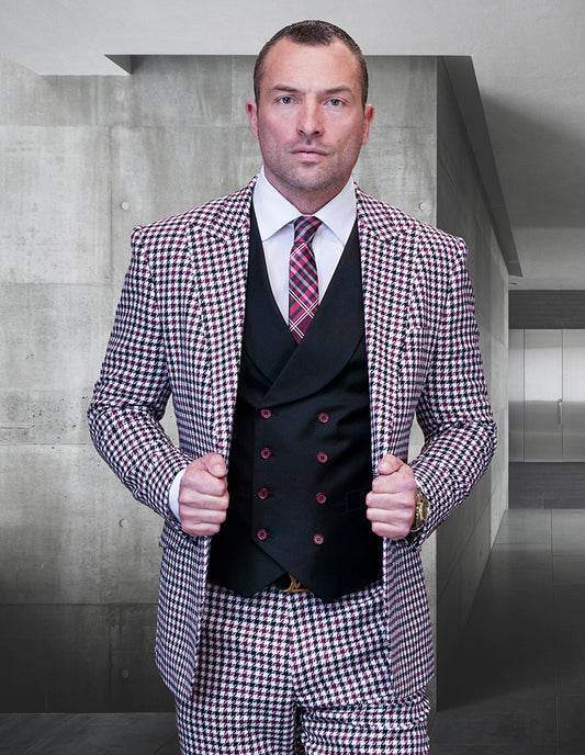 Dressed in the Masimo-Burgundy suit from Statement Clothing, a man stands in a contemporary hallway, confidently adjusting the sleek lapels of his modern-fit houndstooth jacket made from fine Italian wool. He pairs the look with a black vest and a red plaid tie.