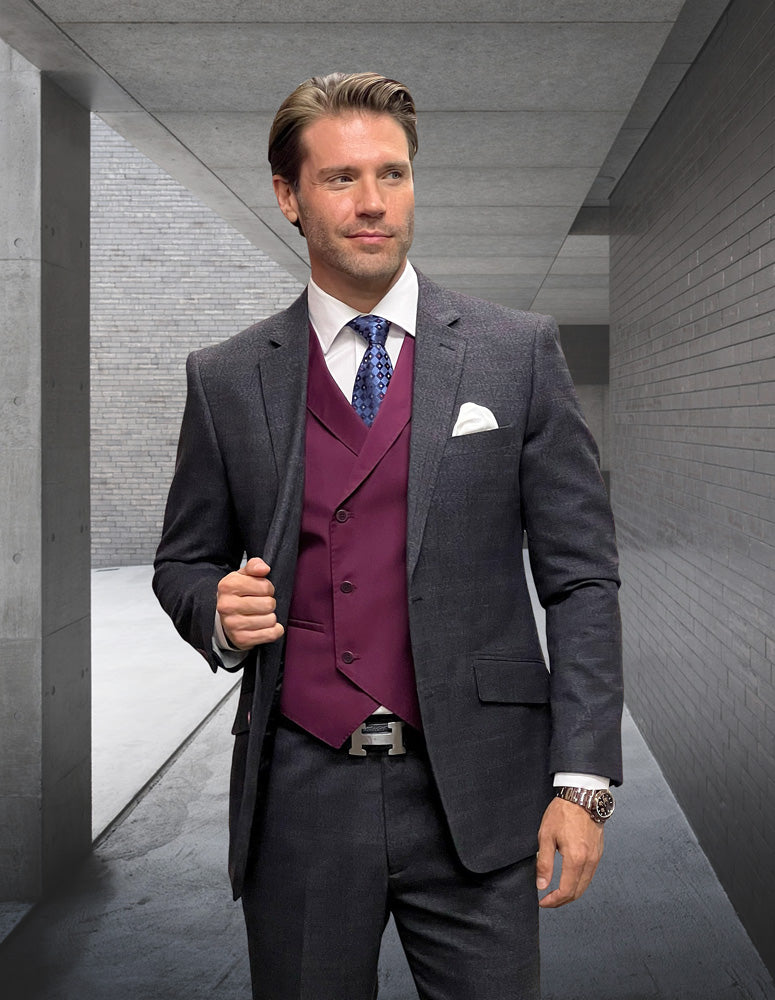 A man in the STATEMENT CLOTHING | MATERA-CHARCOAL suit, featuring a modern-fit and charcoal plaid pattern, complemented by a burgundy vest, stands confidently in a sleek corridor.