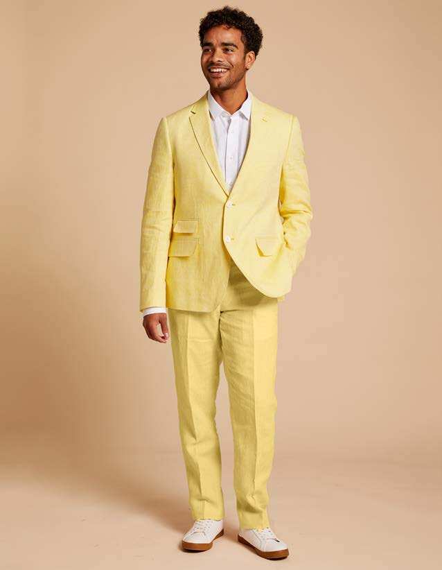 A man in the Inserch Slim Linen Suit SU880-00111 Summer Yellow blazer with a white shirt stands against a beige backdrop, smiling with one hand in his pocket. His slim-fit outfit is perfectly complemented by white sneakers.