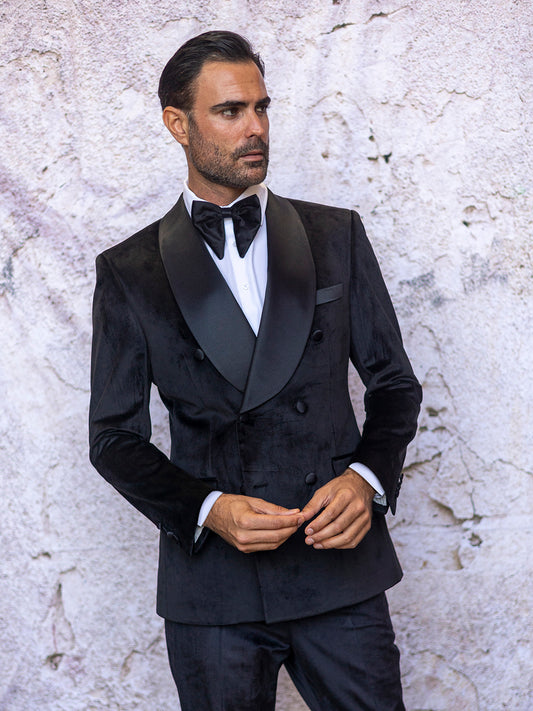 A man in an INSOMNIA MIRAGE 2PC Velvet Tailored Fit Black Suit stands against a textured wall, adjusting his jacket.