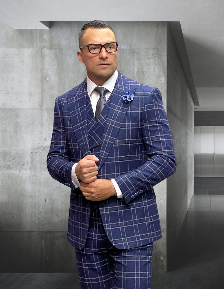 A man in the sleek MONARI-SAPPHIRE suit from Statement Clothing, tailored with luxurious Italian wool in a modern fit and blue plaid design, stands against a concrete wall, exuding sophistication. Glasses perched on his nose enhance the contemporary setting, merging classic style with modern flair.