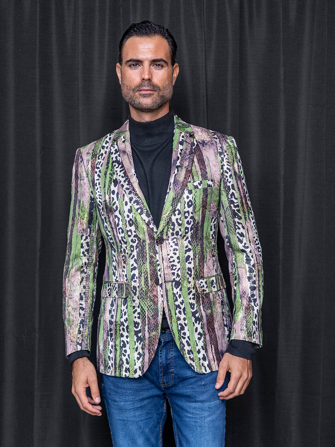 A person stands in front of a dark curtain wearing the INSOMNIA MZS-559 Tailored Fit Green Fancy Sport Coat, perfectly complemented by a black turtleneck and blue jeans.