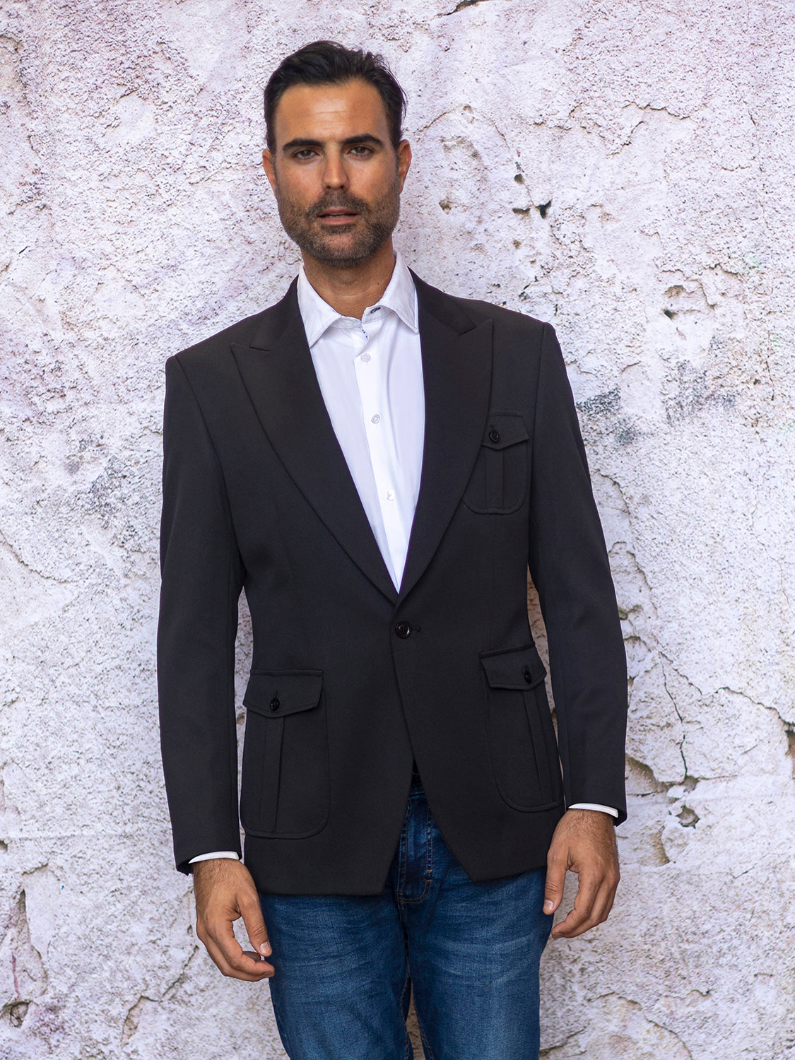 A man in an INSOMNIA MZS-563 tailored fit black fancy sport coat and blue jeans stands against a textured white wall.