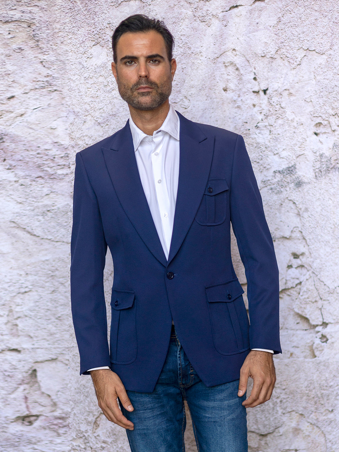 A man in an INSOMNIA MZS-563 Tailored Fit Blue Fancy Sport Coat and a white shirt stands against a textured wall, wearing blue jeans.