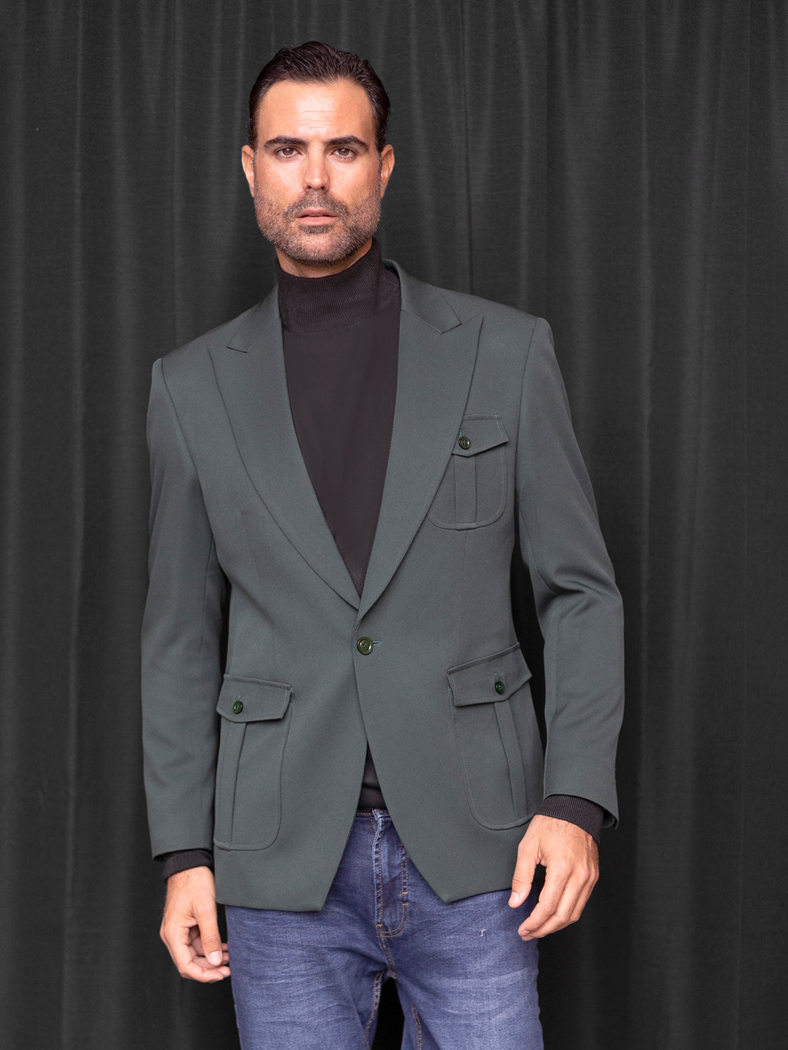 A man with short dark hair stands confidently against a dark curtain backdrop, showcasing the sophisticated INSOMNIA MZS-563 Tailored Fit Hunter Green Fancy Sport Coat from Insomnia, paired elegantly over a black turtleneck and jeans.