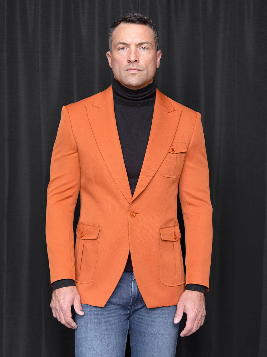 A person wearing the INSOMNIA MZS-563 Tailored Fit Rust Fancy Sport Coat, perfectly paired with a black turtleneck and jeans, stands against a dark backdrop.