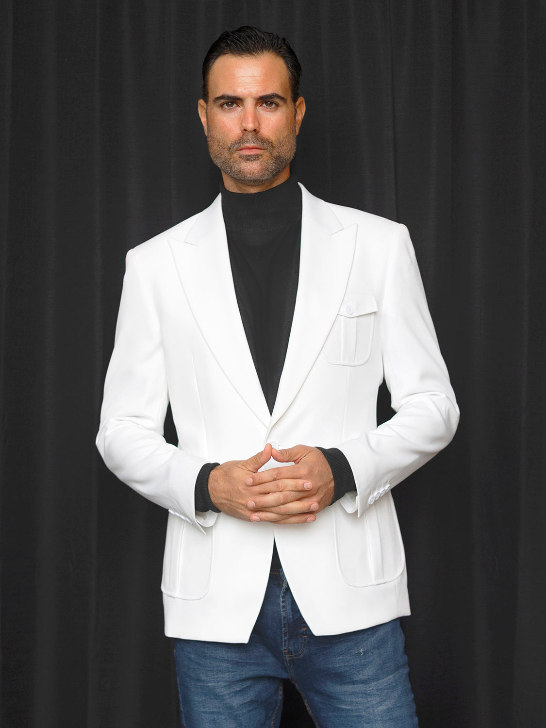 Dressed in the INSOMNIA MZS-563 Tailored Fit White Fancy Sport Coat, a man paired with jeans poses confidently in front of a black curtain, looking at the camera with his hands clasped.