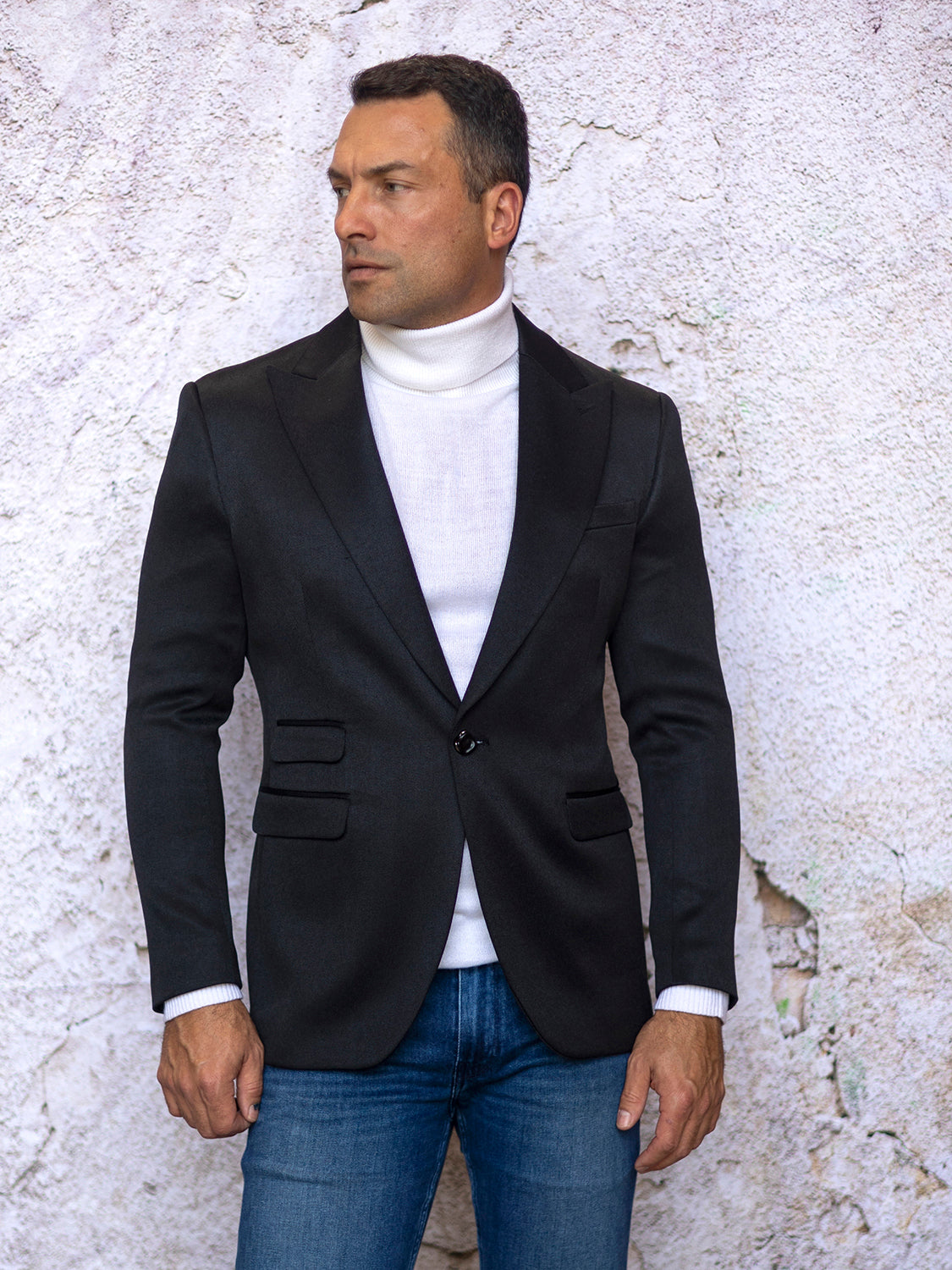 A man wearing the INSOMNIA MZS-564 Tailored Fit Black Fancy Sport Coat and a white turtleneck stands against a textured white wall, gazing to the side.