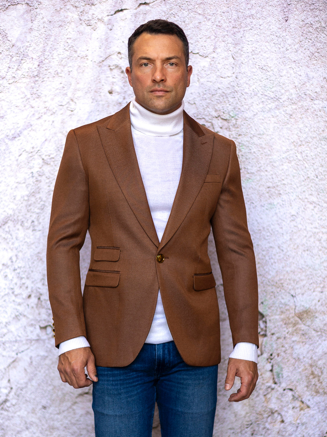 A man in an INSOMNIA MZS-564 Tailored Fit Copper Fancy Sport Coat and a white turtleneck stands against a light textured wall, wearing blue jeans.