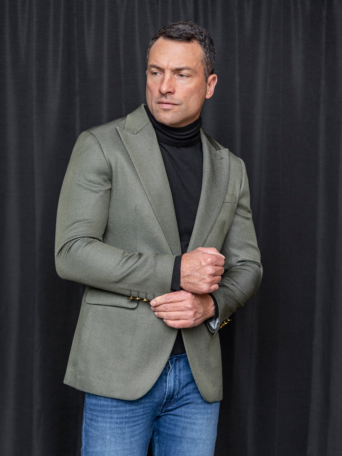 A man wearing the INSOMNIA MZS-564 Tailored Fit Hunter Green Fancy Sport Coat from Insomnia and a black turtleneck stands against a dark curtain, adjusting his sleeve.