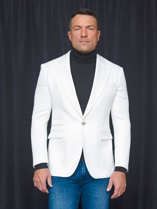 A man stands against a dark curtain, exuding sophistication in his tailored fit ensemble featuring the INSOMNIA MZS-564 Tailored Fit White Fancy Sport Coat, paired with a black turtleneck and blue jeans.