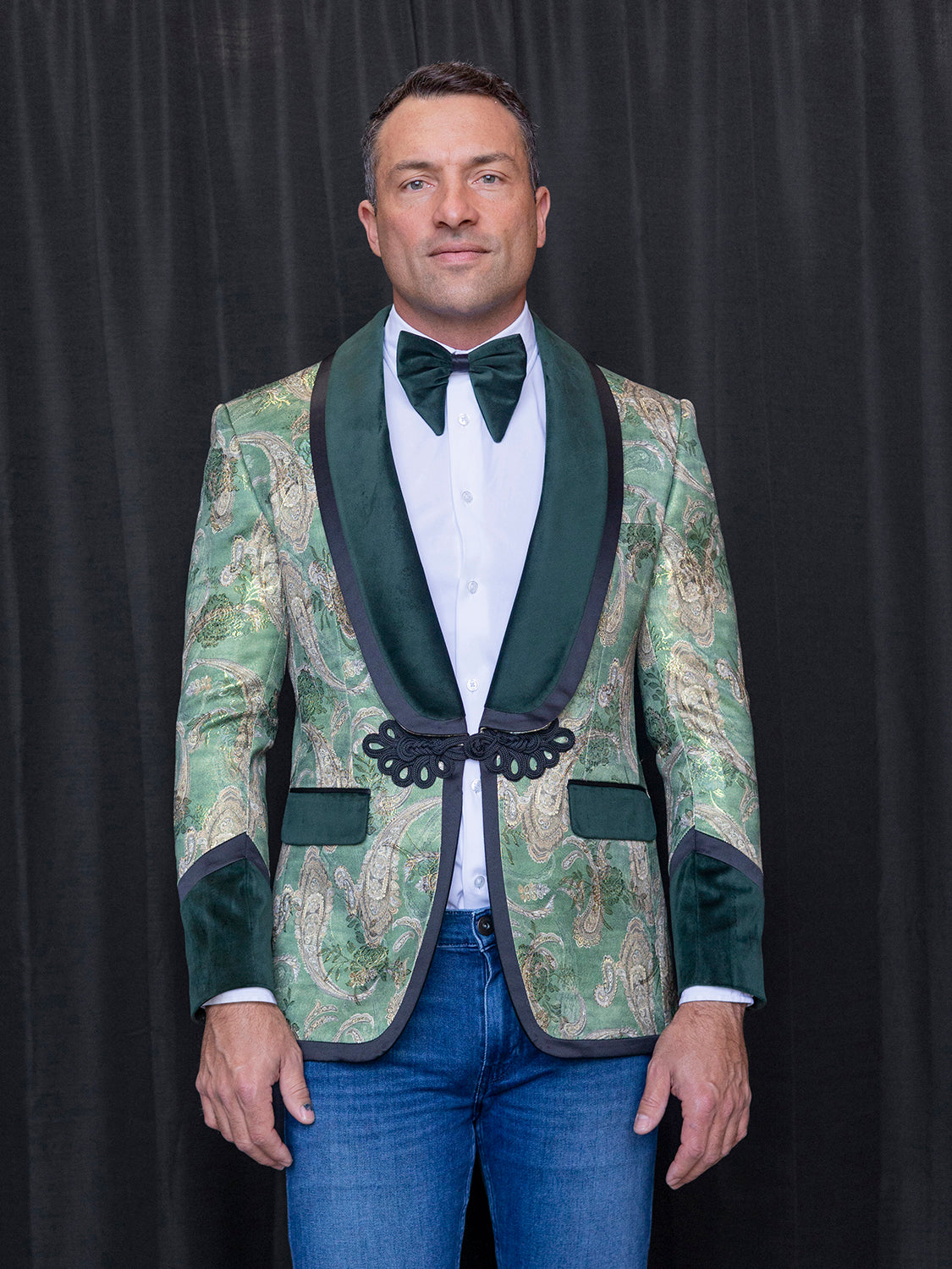 The man is dressed in the INSOMNIA MZS-566 Tailored Fit Green Fancy Sport Coat with black lapels, paired with a white shirt and a black bow tie. His ensemble is completed with blue jeans as he stands against a black background.