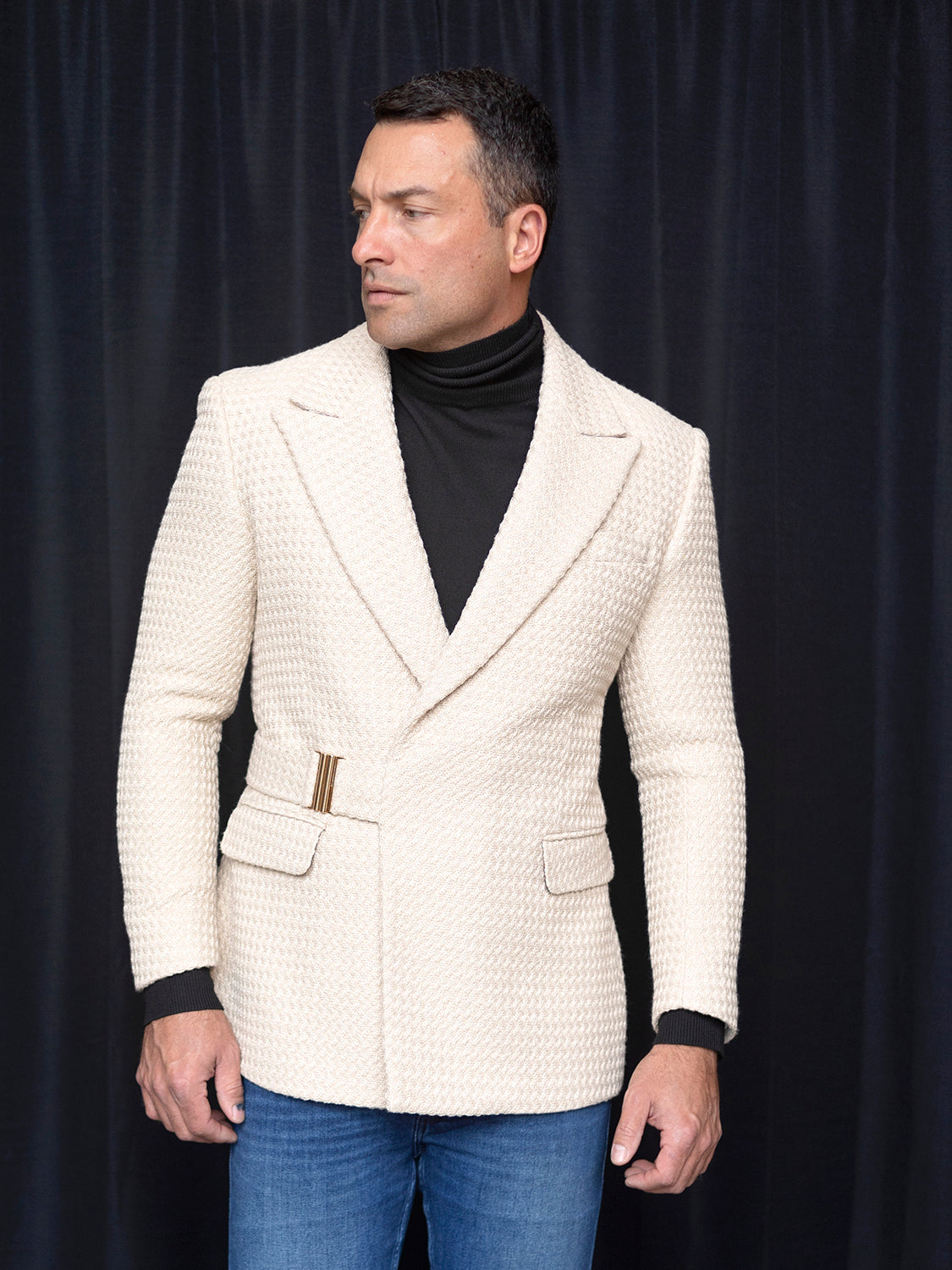 A man in an INSOMNIA MZS-567 Tailored Fit Tan Fancy Sport Coat and black turtleneck stands against a dark curtain, looking to his left.