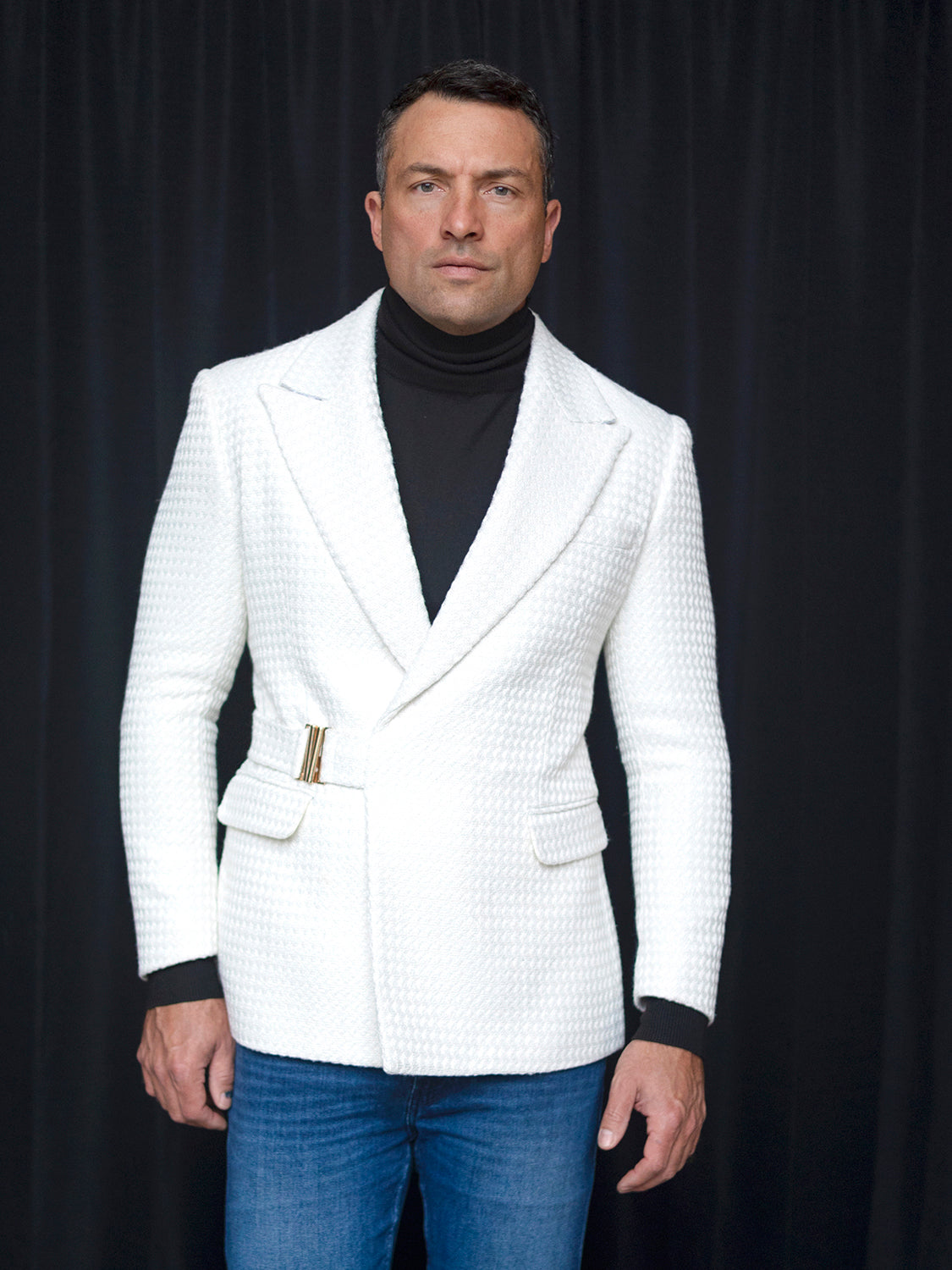 A man stands in front of a black curtain wearing the INSOMNIA MZS-567 Tailored Fit White Fancy Sport Coat, which features a textured finish. It is perfectly paired with a snug black turtleneck and well-fitted blue jeans.