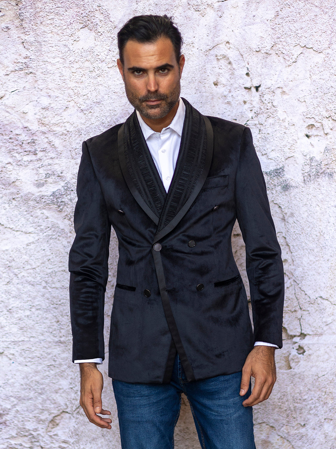 A man wearing the INSOMNIA MZV-562 Tailored Fit Black Fancy Velvet Sport Coat paired with jeans stands against a textured wall, looking at the camera.