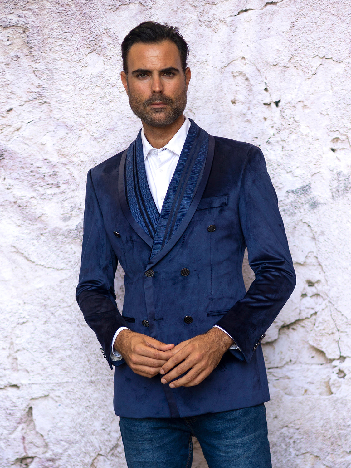 A man with dark hair and a beard confidently stands against a textured white wall, highlighting his tailored fit in an Insomnia INSOMNIA MZV-562 Tailored Fit Blue Fancy Velvet Sport Coat paired with jeans.