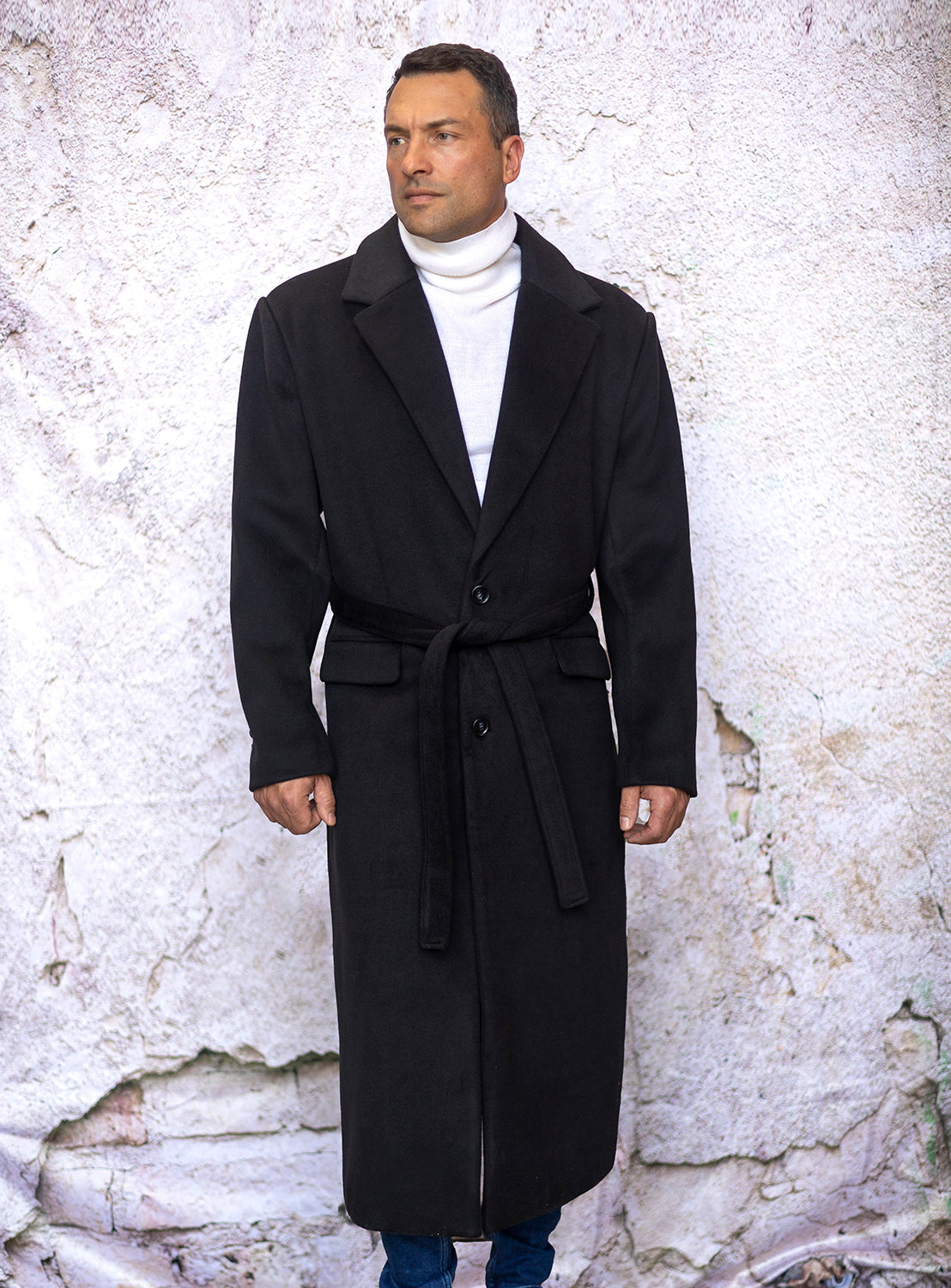 A man wearing an Insomnia INSOMNIA MZW-500 Black Trench Coat and a white turtleneck stands against a textured, light-colored wall.