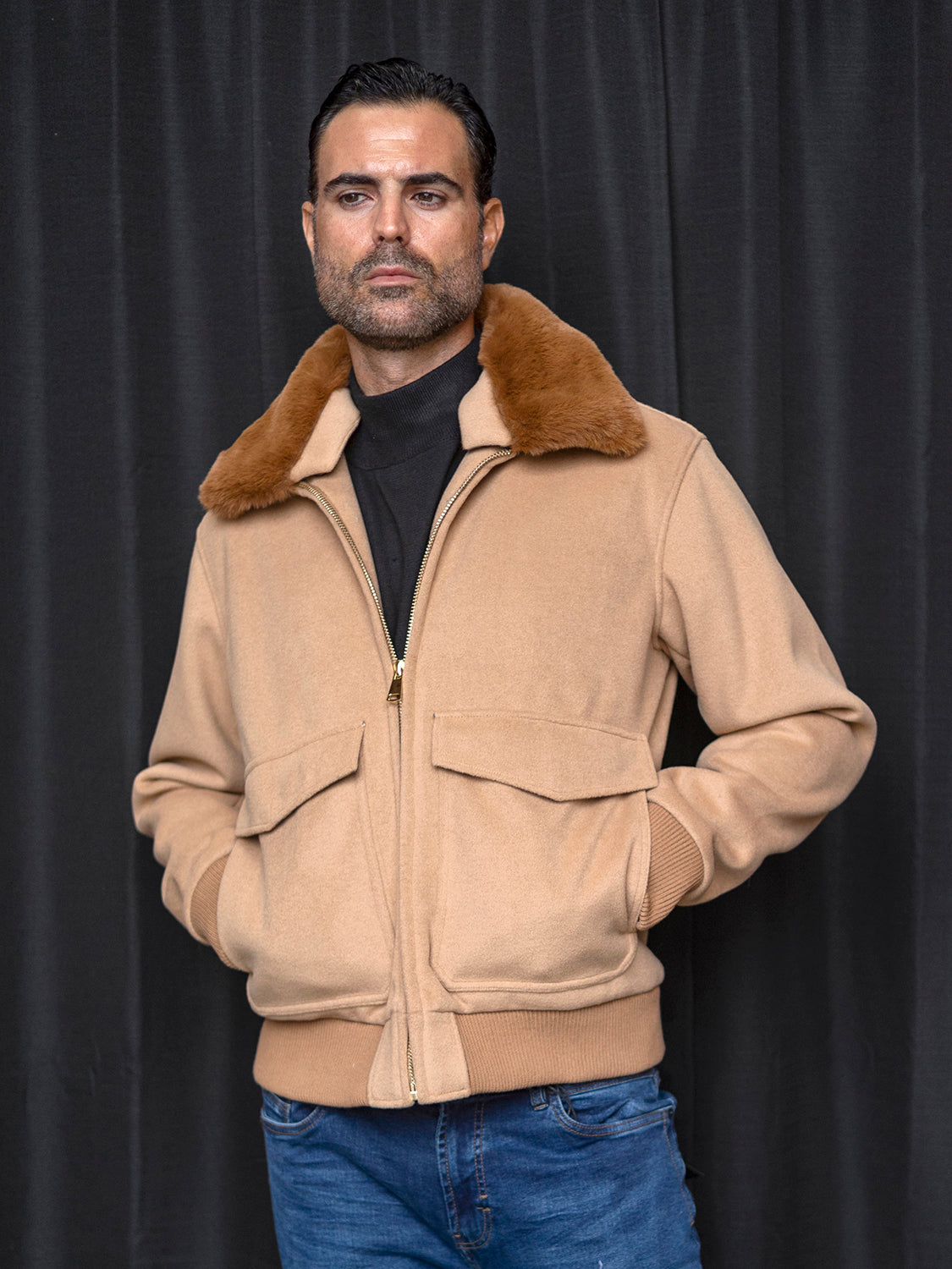 A man wearing the INSOMNIA MZW-501 Tailored Fit Camel Wool Bomber Jacket stands with hands in pockets against a dark backdrop, complemented by his blue jeans.